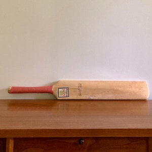 May include: A wooden cricket bat with a pink grip and a blue and white label. The bat is lying on a wooden table.