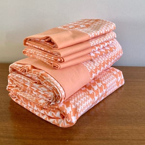 May include: Neatly folded peach-coloured bed sheets with a white houndstooth pattern, stacked on a wooden surface. The top sheet has a solid peach border. The sheets are arranged to display the fabric's design and colour.