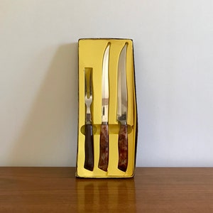 Può includere: Un set di tre coltelli da cucina con manici marmorei marroni in una scatola gialla. I coltelli sono un coltello da intaglio, un coltello per bistecche e una forchetta.