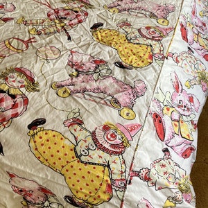 May include: A white cotton bed sheet with a repeating pattern of clowns, bunnies, and dogs. The clowns are wearing yellow and red outfits with polka dots. The bunnies are pink and white. The dogs are pink and have black eyes. The sheet is in good condition and has a vintage look.