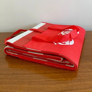 May include: A red and white foldable shopping bag with a white logo on the front. The bag is folded and sitting on a wooden surface.