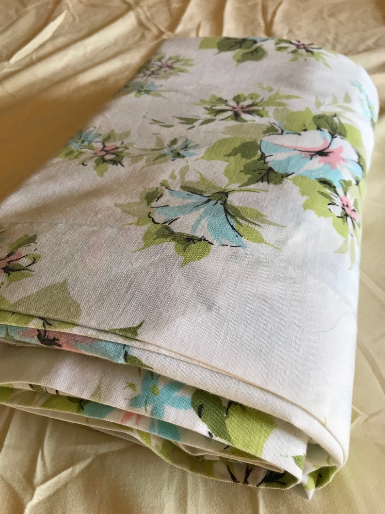 Puede incluir: Una s&aacute;bana de tela blanca con un patr&oacute;n floral en tonos de verde, azul y rosa. La s&aacute;bana est&aacute; doblada y tiene una superficie suave y texturizada.