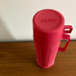 May include: A pink thermos with a handle and the word "THERMOS" embossed on the lid.