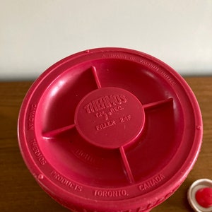 May include: Red thermos bottle with a white lid. The bottom of the bottle has the text "CANADIAN THERMOS A BRAND OF VACUUM BOTTLE THERMOS T.M. REG. FILLER 24F PRODUCTS TORONTO, CANADA".