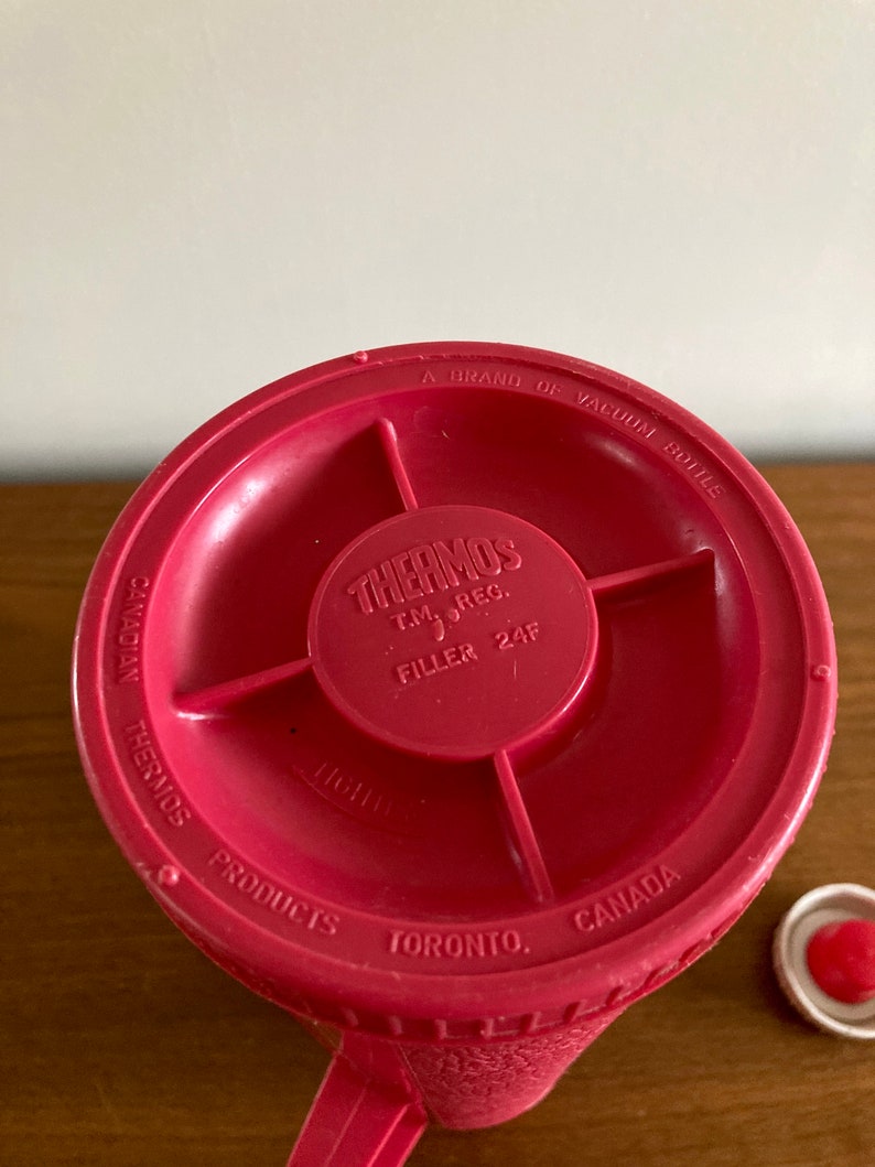May include: Red thermos bottle with a white lid. The bottom of the bottle has the text "CANADIAN THERMOS A BRAND OF VACUUM BOTTLE THERMOS T.M. REG. FILLER 24F PRODUCTS TORONTO, CANADA".