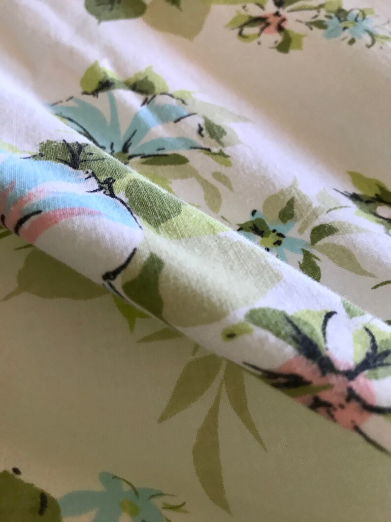 Puede incluir: Primer plano de una tela blanca con un patr&oacute;n floral verde y azul. La tela est&aacute; doblada y el patr&oacute;n es visible en ambos lados del pliegue.