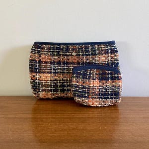 May include: Two fabric cosmetic bags with a black and orange plaid pattern. The larger bag is lying flat and the smaller bag is standing upright in front of it.
