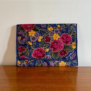 May include: A blue floral patterned clutch with a gold trim and the text "Tumi" in white.