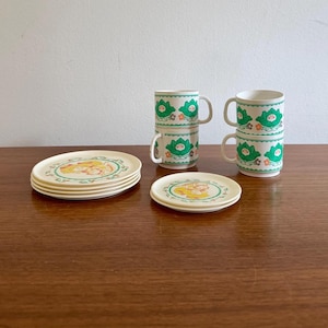 May include: A vintage tea set featuring cream-coloured plates and mugs. The plates have a cartoon character design with a green border. The mugs are white with a green floral pattern. The set includes four plates and four mugs.