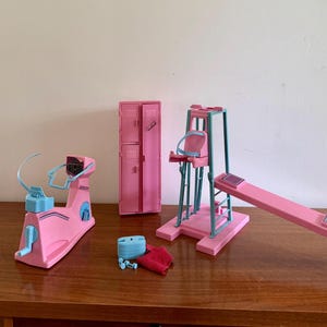 Op de afbeelding: Een verzameling roze en blauwe miniatuur fitnessapparatuur, waaronder een hometrainer, een gewichtmachine en een halterbank. Ook inbegrepen zijn een roze locker en accessoires.