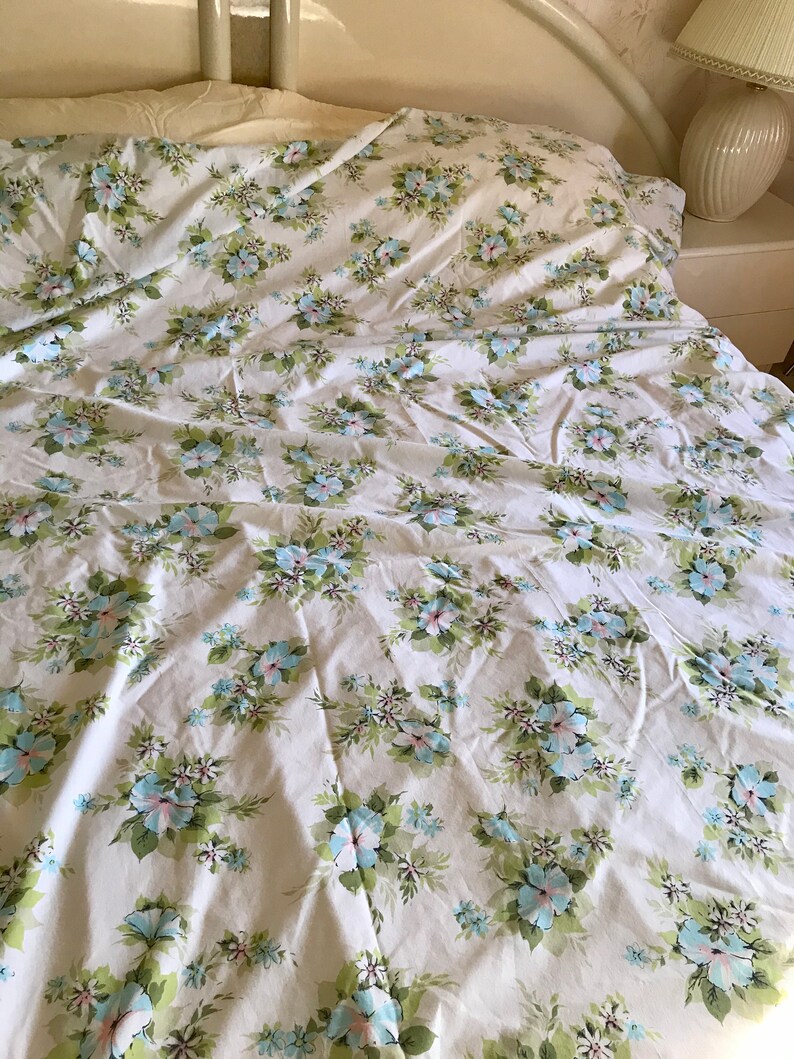 Puede incluir: S&aacute;banas blancas de cama con un patr&oacute;n floral con flores azules y rosas sobre un fondo blanco.