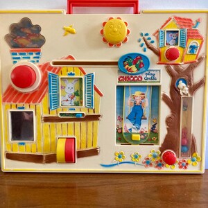 Puede incluir: Un juguete infantil vintage con una base color crema, un asa roja y varios elementos interactivos. Incluye una casa con un conejo, un &aacute;rbol con un rat&oacute;n, un sol y el texto "CHICCO play center".