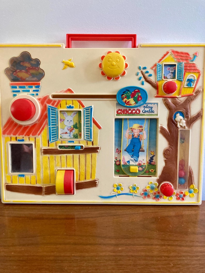 Puede incluir: Un juguete infantil vintage con una base color crema, un asa roja y varios elementos interactivos. Incluye una casa con un conejo, un &aacute;rbol con un rat&oacute;n, un sol y el texto "CHICCO play center".