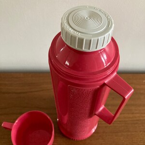 May include: A pink thermos with a white lid and a matching pink cup. The lid has the words "To Tighten" and "To Remove" printed on it.