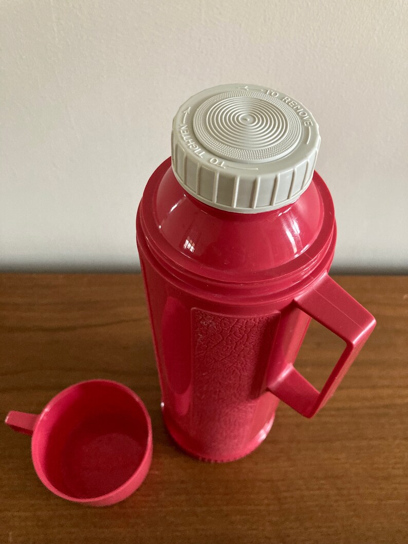 May include: A pink thermos with a white lid and a matching pink cup. The lid has the words "To Tighten" and "To Remove" printed on it.