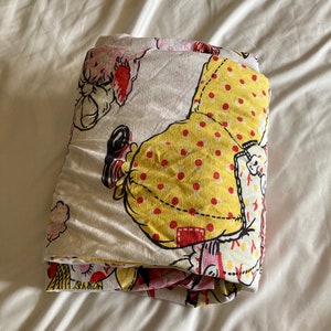 May include: Folded white fabric with a yellow, red, and pink print of a cartoon doll. The doll is wearing a yellow dress with red polka dots and a red hat.