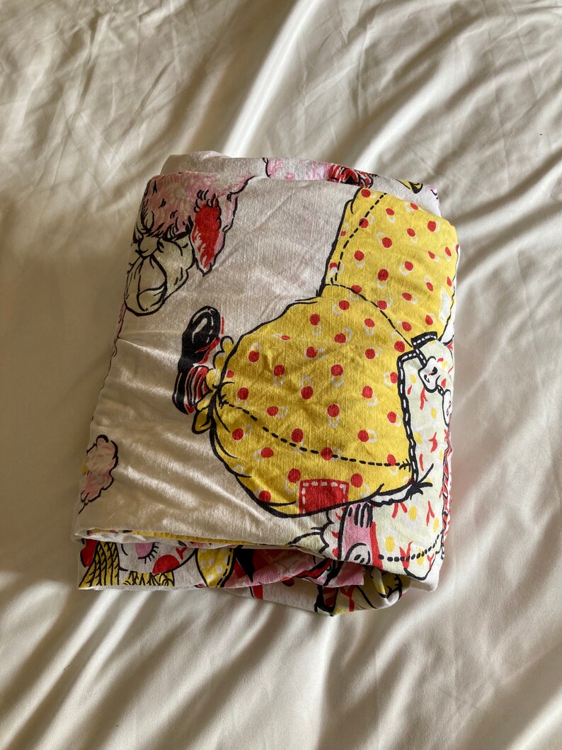 May include: Folded white fabric with a yellow, red, and pink print of a cartoon doll. The doll is wearing a yellow dress with red polka dots and a red hat.