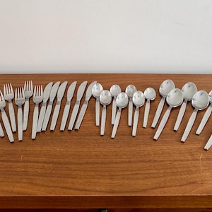 Puede incluir: Un juego de cubiertos de acero inoxidable, que incluye tenedores, cuchillos y cucharas, dispuestos sobre una superficie de madera. La cubertería tiene un diseño sencillo y moderno con acabado plateado y mangos blancos. El juego incluye cucharas de varios tamaños.