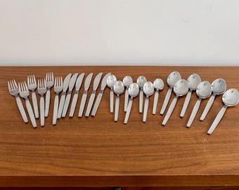 Op de afbeelding: Een verzameling roestvrijstalen bestek, waaronder vorken, messen en lepels, gerangschikt op een houten oppervlak. Het zilverwerk heeft een eenvoudig, modern ontwerp met een zilveren afwerking en witte handvatten. De set bevat verschillende maten lepels.