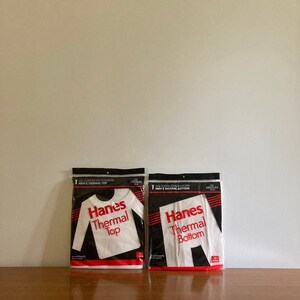 Peut inclure: Deux paquets de sous-v&ecirc;tements thermiques blancs, un haut et un bas, tous deux portant l'inscription "Hanes Thermal" en lettres rouges. Les paquets sont sur une surface en bois.