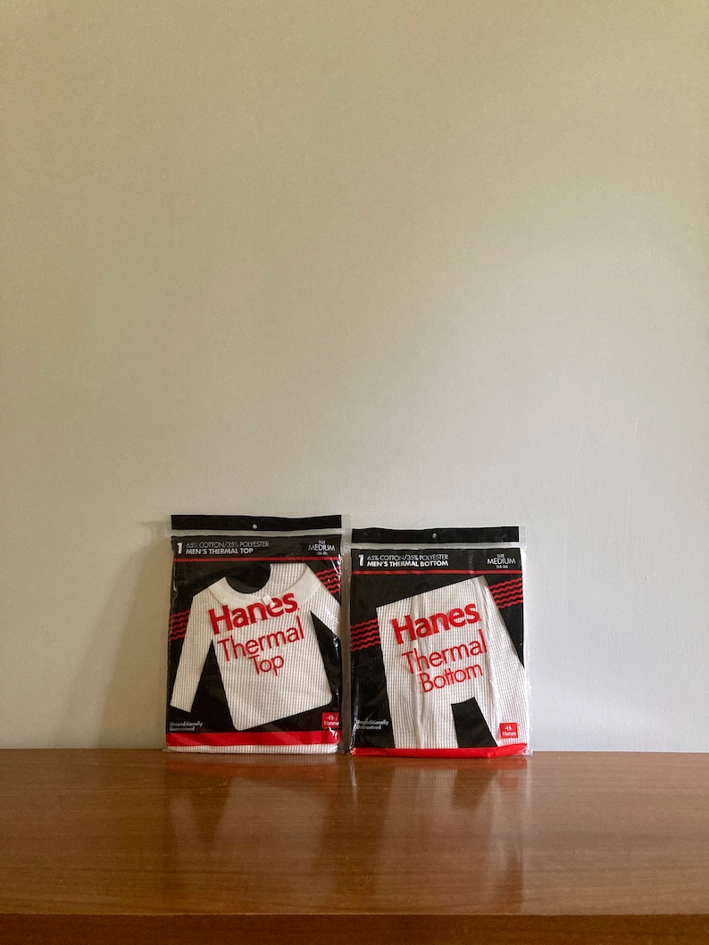Peut inclure: Deux paquets de sous-v&ecirc;tements thermiques blancs, un haut et un bas, tous deux portant l'inscription "Hanes Thermal" en lettres rouges. Les paquets sont sur une surface en bois.