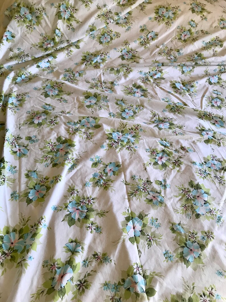 Puede incluir: S&aacute;bana blanca de cama con un patr&oacute;n floral con flores azules y rosas sobre tallos verdes. La s&aacute;bana est&aacute; arrugada y tiene un aspecto vintage.