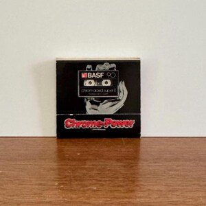 May include: A black cassette tape box with the text "BASF 90" and "Chrome-Power" in red and white. The box features a graphic of a cassette tape held by a hand. The background is white and the box is on a wooden surface.