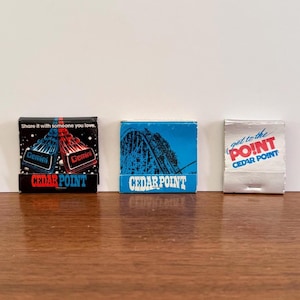 May include: Three vintage matchbooks from Cedar Point amusement park. One matchbook has a black background with the word "Gemini" and "CEDAR POINT". Another has a blue background with a roller coaster and "CEDAR POINT". The third is white with "get to the POINT" and "CEDAR POINT".