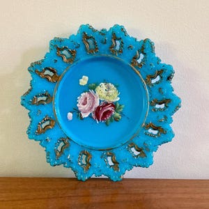 May include: A vintage, turquoise-colored decorative plate with a scalloped edge and gold accents. The center features a hand-painted floral design with pink, red, and yellow roses. The plate is likely made of glass.