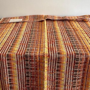 May include: A woven textile with a striped pattern in shades of orange, yellow, brown, and white. The fabric appears to be folded over at the top, with a small label visible. The textile is likely a blanket or throw.