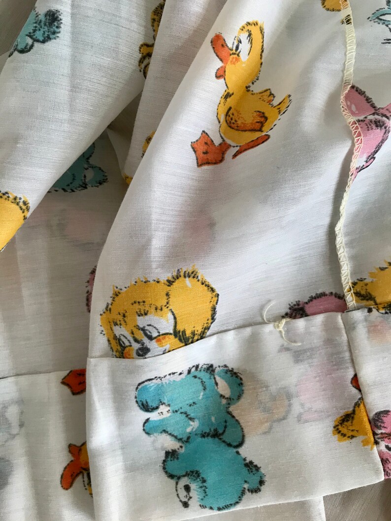 May include: White fabric with a repeating pattern of cartoon ducks and bears. The ducks are yellow with orange beaks and feet. The bears are blue, pink, and yellow.