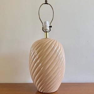 Könnte beinhalten: Eine rosa Keramik-Tischlampe mit einem gedrehten Design. Die Lampe hat einen goldenen Metallbogen und eine weiße Fassung.