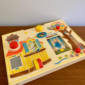 Puede incluir: Un juguete infantil vintage con una base color crema y detalles coloridos. El juguete presenta un dise&ntilde;o de casa con un conejito, un ni&ntilde;o y varios elementos interactivos como botones y deslizadores. El texto "Chicco Castle" es visible.