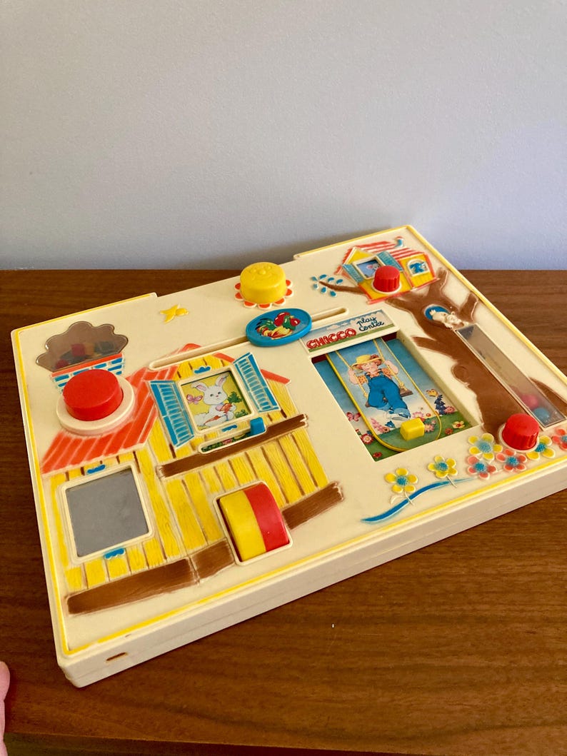 Puede incluir: Un juguete infantil vintage con una base color crema y detalles coloridos. El juguete presenta un dise&ntilde;o de casa con un conejito, un ni&ntilde;o y varios elementos interactivos como botones y deslizadores. El texto "Chicco Castle" es visible.
