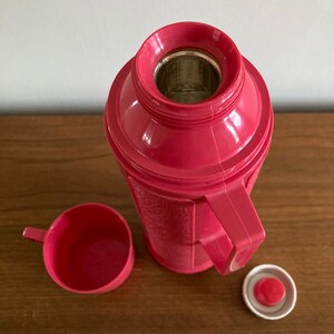 May include: A pink thermos with a matching pink cup and lid. The thermos has a textured surface and a handle.