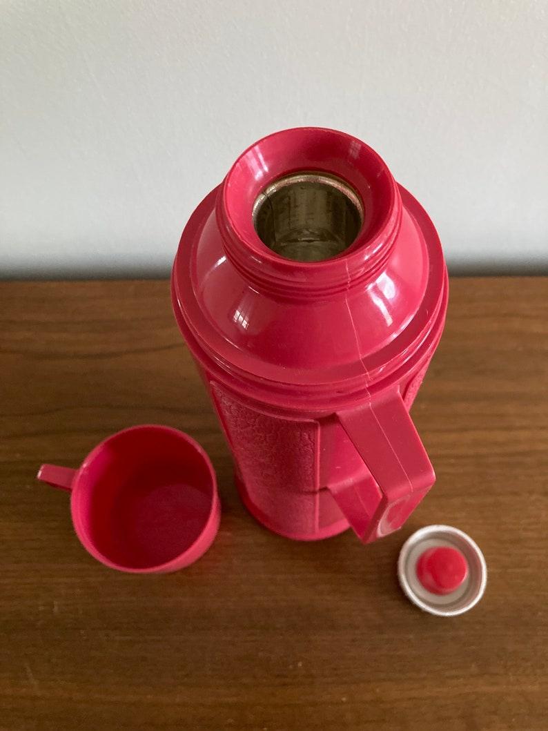 May include: A pink thermos with a matching pink cup and lid. The thermos has a textured surface and a handle.