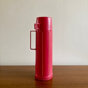 May include: A vintage pink thermos with a carrying handle. The thermos has a textured surface and a silver lid.