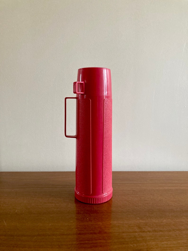 May include: A vintage pink thermos with a carrying handle. The thermos has a textured surface and a silver lid.