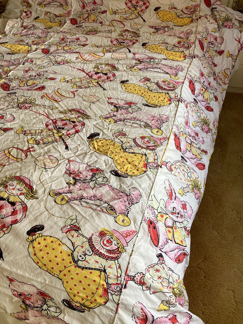 May include: A white cotton bed sheet with a repeating pattern of clowns, bunnies, and dogs. The clowns are wearing yellow and red outfits with polka dots. The bunnies are pink and white. The dogs are pink and have black eyes. The sheet is in good condition and has a vintage look.
