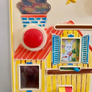 Puede incluir: Una colorida casa de juguete vintage con techo rojo, paredes amarillas y contraventanas azules. El juguete presenta un bot&oacute;n rojo, una chimenea con una escena de nubes y una ventana con un conejito sosteniendo una zanahoria. Tambi&eacute;n se ve un p&aacute;jaro amarillo.