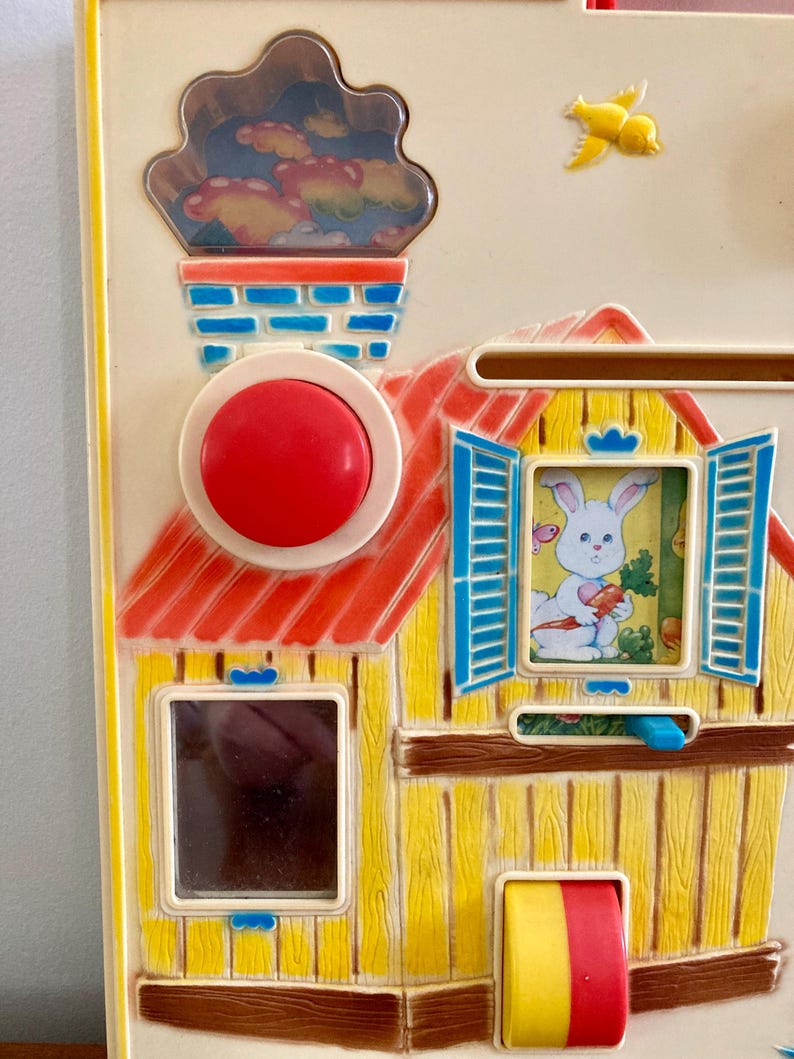 Puede incluir: Una colorida casa de juguete vintage con techo rojo, paredes amarillas y contraventanas azules. El juguete presenta un bot&oacute;n rojo, una chimenea con una escena de nubes y una ventana con un conejito sosteniendo una zanahoria. Tambi&eacute;n se ve un p&aacute;jaro amarillo.