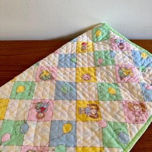 Puede incluir: Una manta de bebé cuadrada y acolchada con un diseño de patchwork. Cada cuadrado presenta un personaje de Cabbage Patch Kids con globos. La manta tiene un borde verde y un patrón de cuadros.