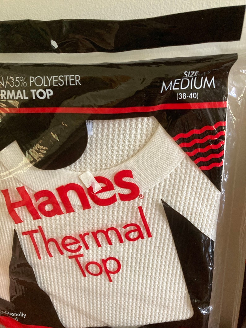 Peut inclure: Un haut thermique blanc Hanes dans un sac en plastique. Le sac porte le texte "35% Polyester Thermal Top" et "Taille M (38-40)".
