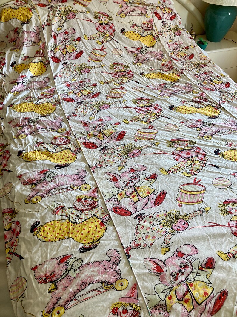 May include: A white cotton fabric with a repeating pattern of pink and yellow toy animals. The animals include a clown, a bunny, and a cat. The animals are all wearing clothes and have a whimsical, vintage look.