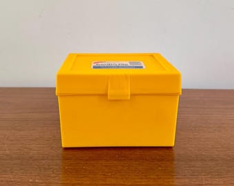 May include: A bright yellow plastic box with a lid and a clasp closure. The box has a rectangular shape and a label on the top that reads "Jumbo File". The box is sitting on a wooden surface.