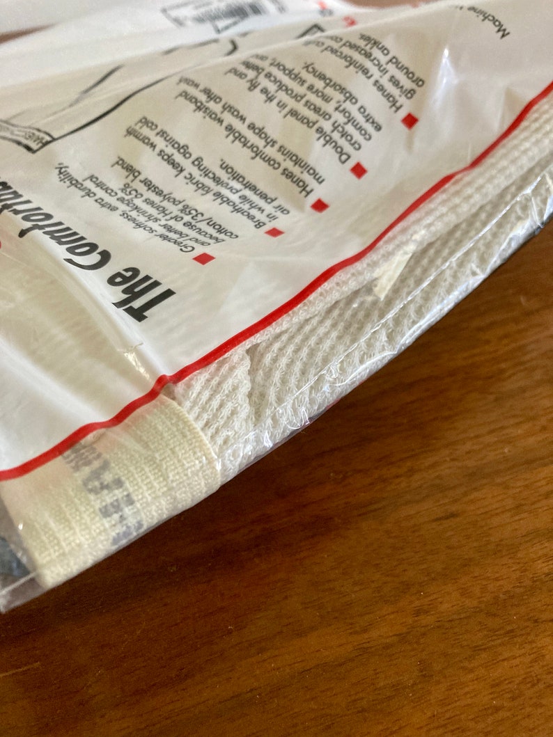 Peut inclure: Un paquet blanc avec du texte rouge qui indique "The Comforta" et "Hanes 65% coton/35% polyester blend." Le paquet contient un produit en coton blanc.