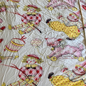 May include: A white fabric with a repeating pattern of red, yellow, and pink clowns, dolls, and toy animals. The clowns are wearing yellow and red outfits with polka dots and buttons. The dolls are wearing pink and red plaid dresses. The toy animals are pink and white.