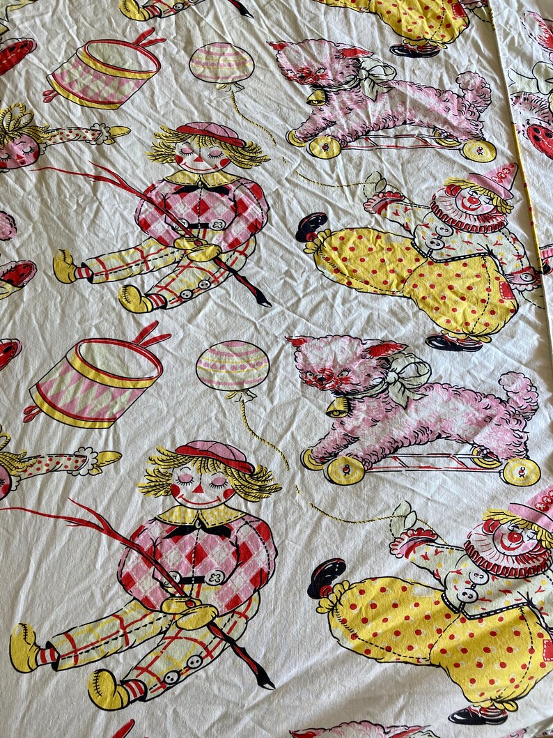 May include: A white fabric with a repeating pattern of red, yellow, and pink clowns, dolls, and toy animals. The clowns are wearing yellow and red outfits with polka dots and buttons. The dolls are wearing pink and red plaid dresses. The toy animals are pink and white.