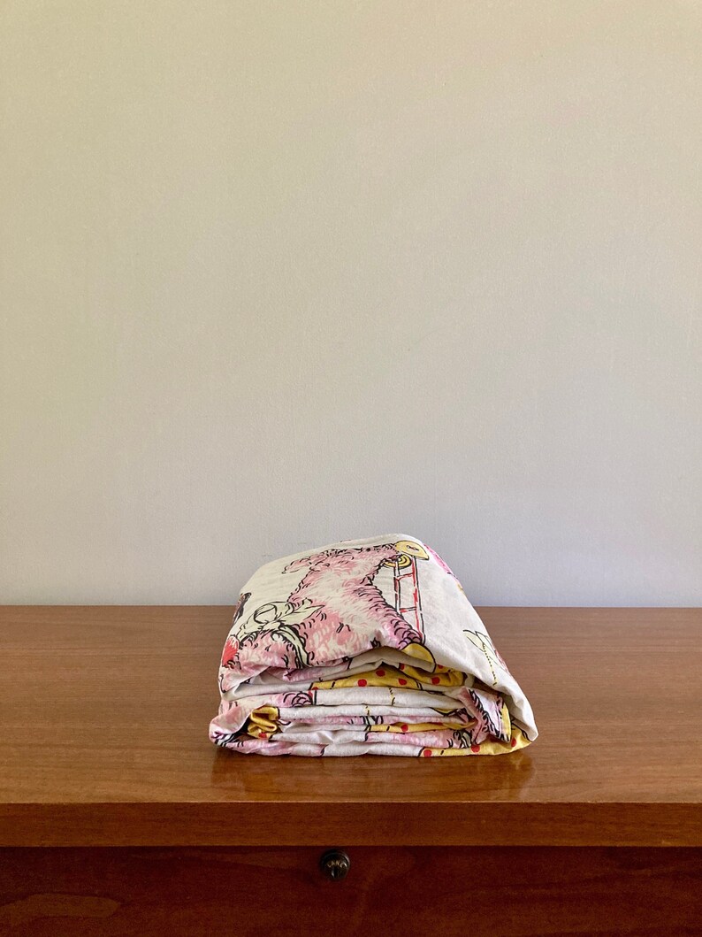 May include: A folded stack of fabric with a pink, yellow, and white pattern featuring cartoon animals. The fabric is folded on a wooden surface.
