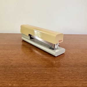May include: A vintage Bates stapler in cream and silver. The stapler has a rectangular shape with a cream-colored top and a silver base. The word "Bates" is printed in red on the side. The stapler is sitting on a wooden surface.